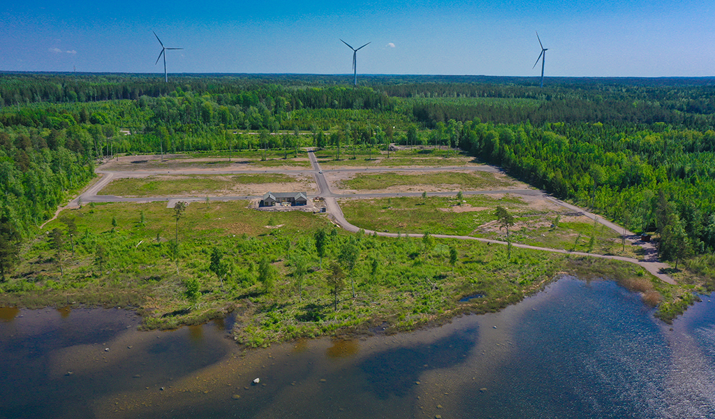 Drönarbild på tomtområde, vatten och skog. I bakgrunden syns tre stycken vindkraftverk