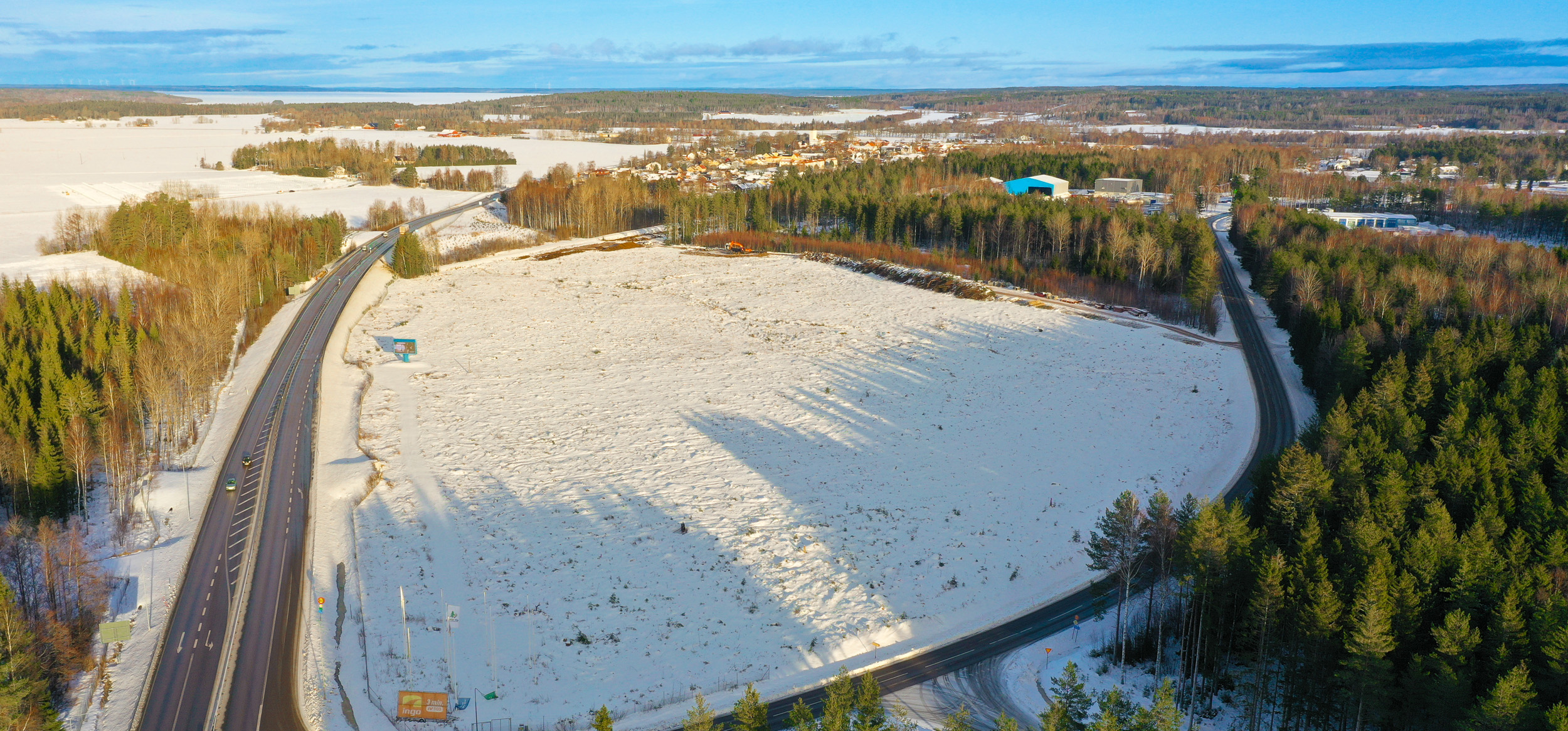 drönarbild över etableringsområde. Vinterlandskap, två vägar och skog, samt en horisont med blå himmel